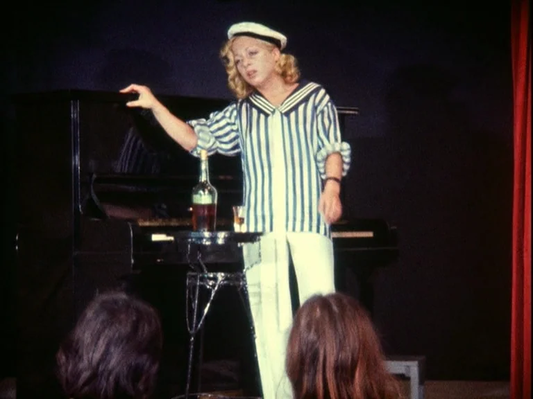 Der Bomberpilot (1970) movie still: medium — A cabaret performer stands on a dimly lit dark wooden floor. She gestures toward a small table with a…