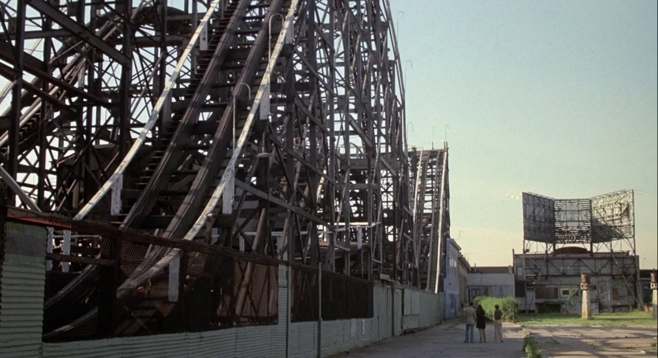 Annie Hall (1977) movie still: wide — A massive wooden roller coaster structure looms over a vacant dirt lot. Three people stand in the lower right…