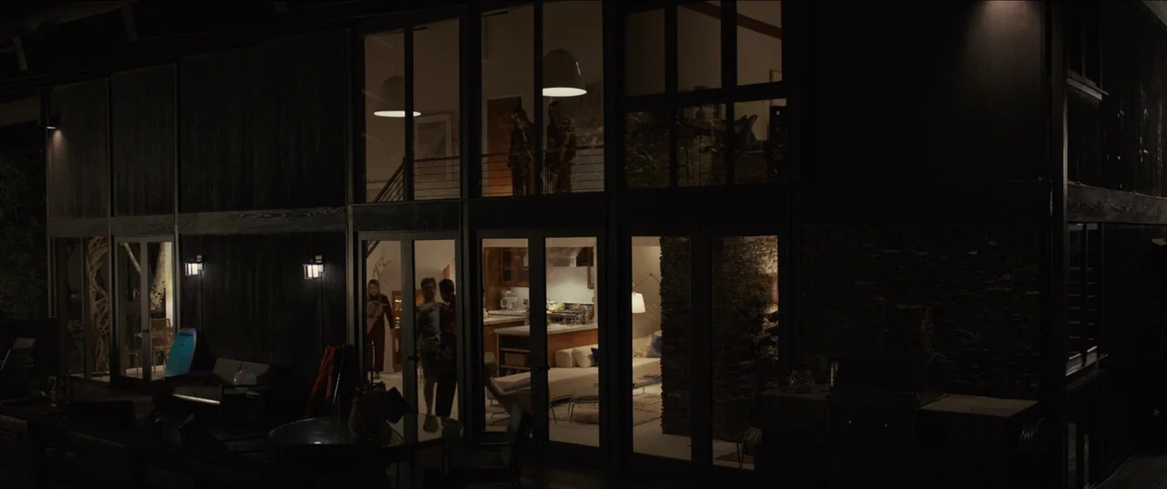 Us (2019) movie still: wide — A dark, modern two-story house features large glass walls and black wooden siding at night. Multiple people move…