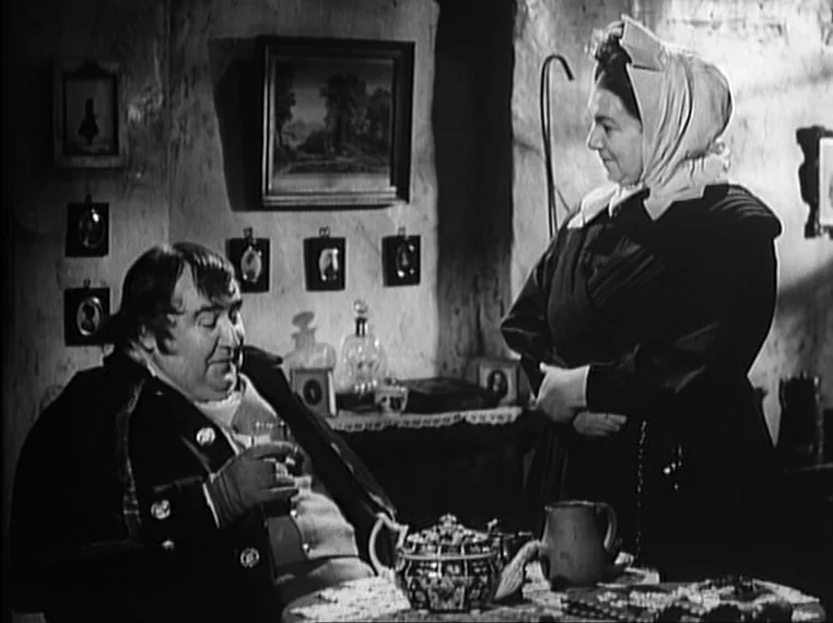 Oliver Twist (1948) movie still: medium — A rustic cottage interior features textured walls adorned with framed pictures and a teapot. A man sits at…