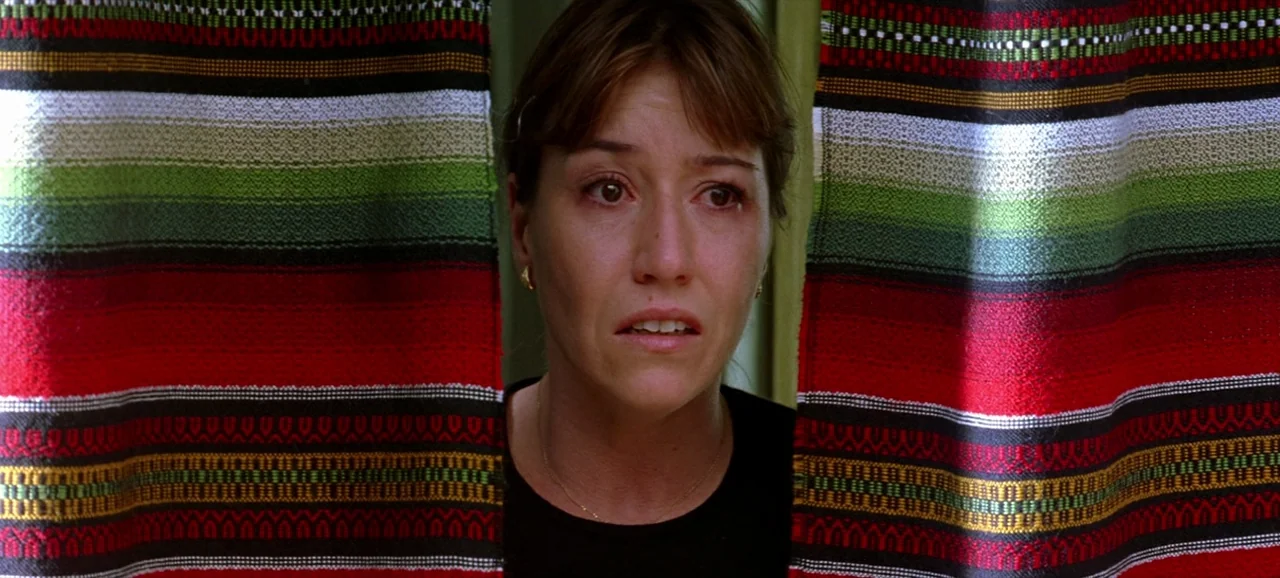 Volver (2006) movie still: closeup — A woman stands behind two panels of vibrant, horizontally striped fabric curtains. She looks directly forward…