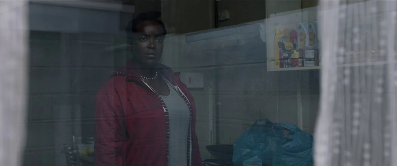 His House (2020) movie still: medium — A dimly lit room with cinder block walls and a high shelf holding groceries. A young woman stands looking…