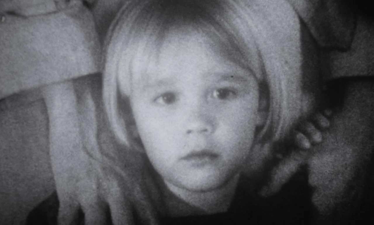 The Secret Son (1979) movie still: closeup — A close portrait of a young child in a sparsely lit interior space. The child faces the camera directly…