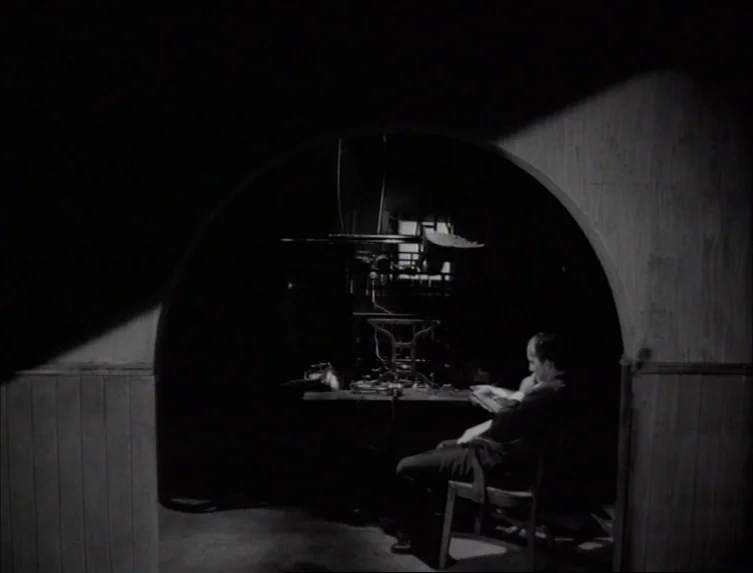 Eraserhead (1977) movie still: medium — A dark subterranean space framed by a stone archway with light hitting the center. A man sits at a small…