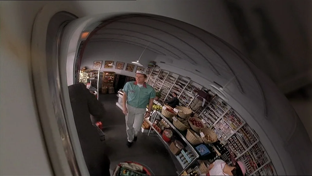 The Truman Show movie still: wide — A small market aisle is reflected in a curved convex security mirror. A store clerk in a uniform walks down the…