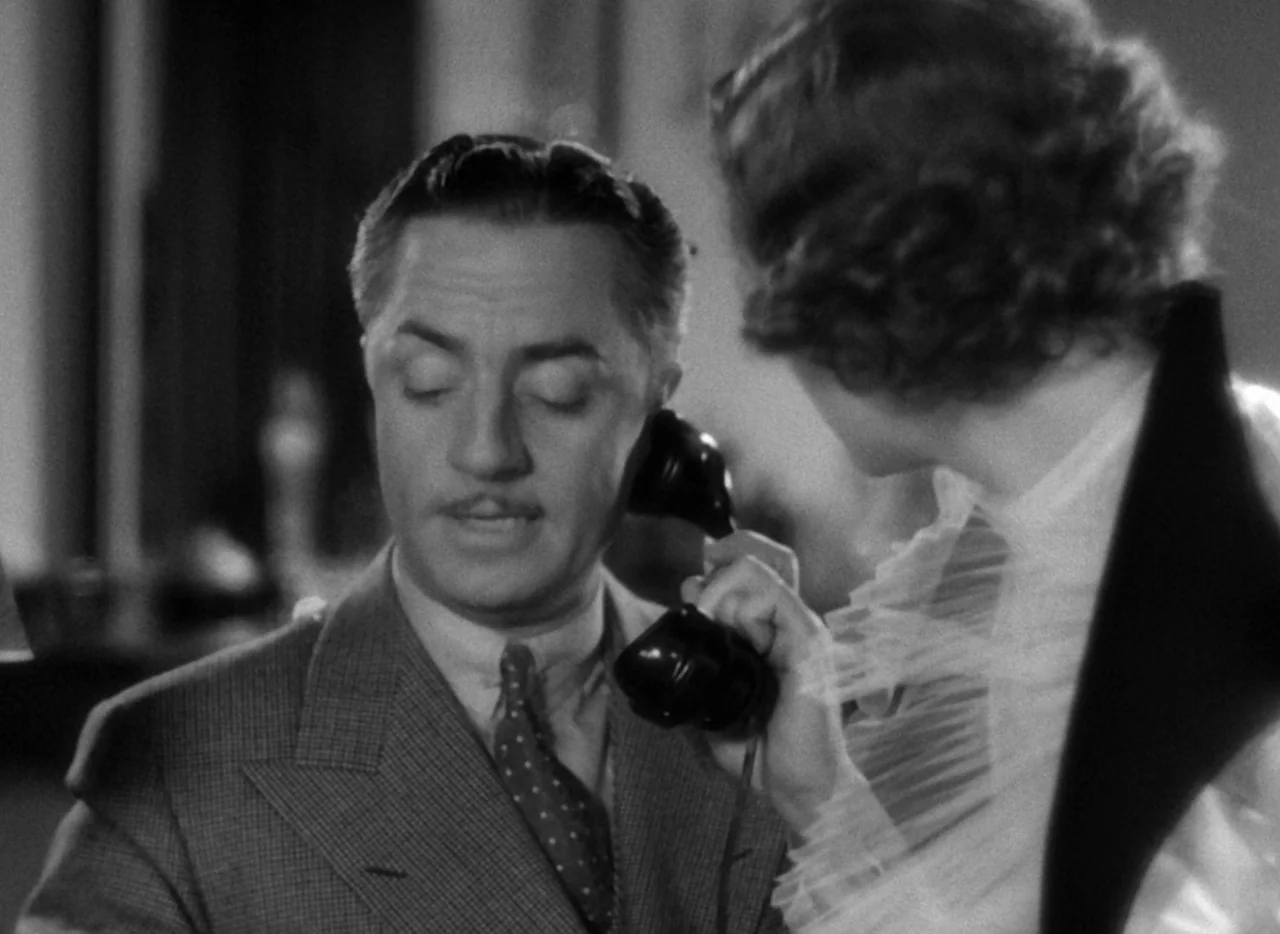 The Thin Man (1934) movie still: medium — A dimly visible interior space features a man and woman in formal attire. A woman reaches forward to place…