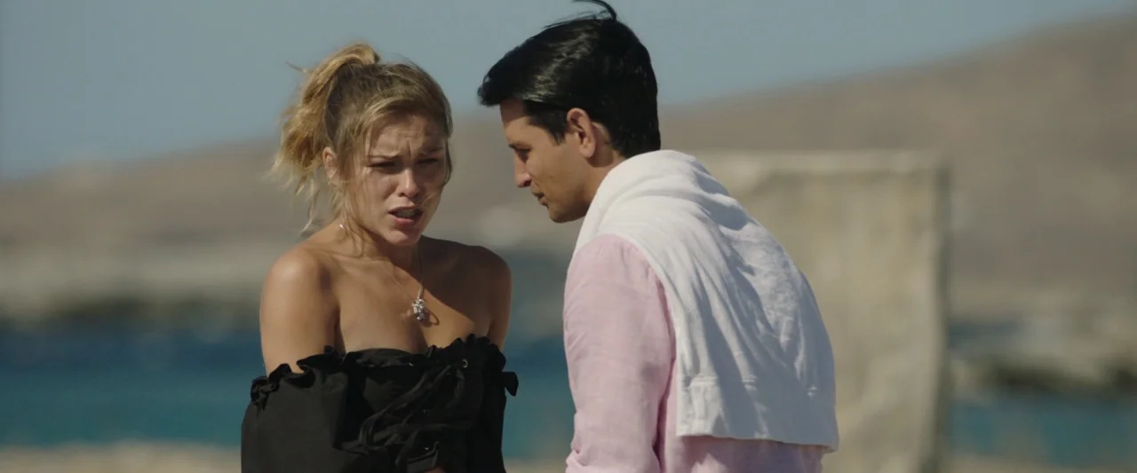 Greed (2019) movie still: medium — A woman and man stand near a shoreline with blurred blue water behind them. They engage in a conversation while…