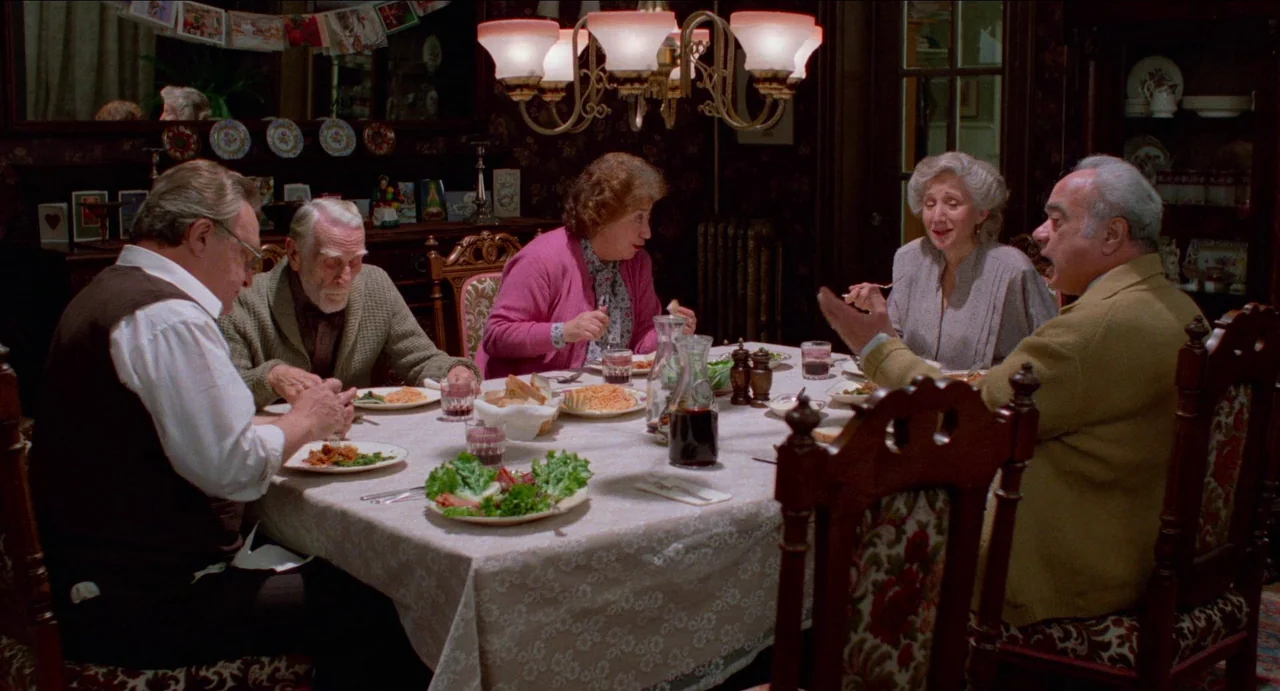 Moonstruck (1987) movie still: wide — A traditional dining room with dark floral wallpaper and wood trim. Five older adults are gathered around a…