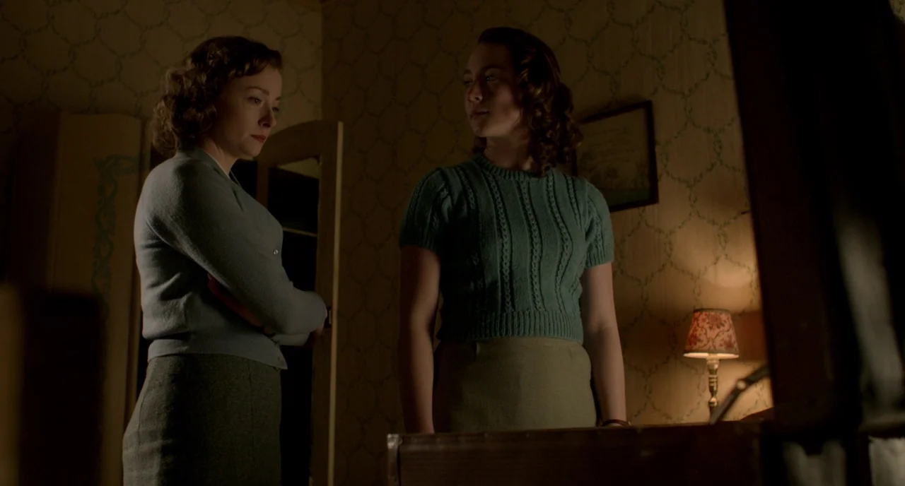 Brooklyn (2015) movie still: medium — An interior bedroom with patterned floral wallpaper and a dim lamp lit at night. Two women in mid-century…
