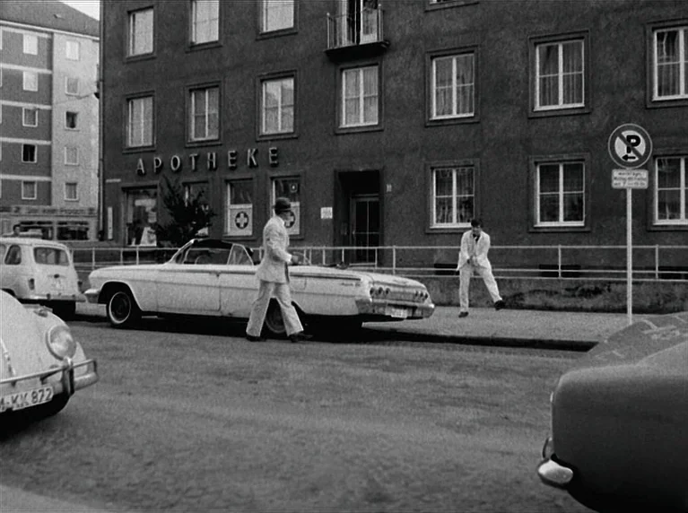 The American Soldier (1970) movie still: wide — A German city street features a multi-story apartment building with an Apotheke sign. Two men walk…