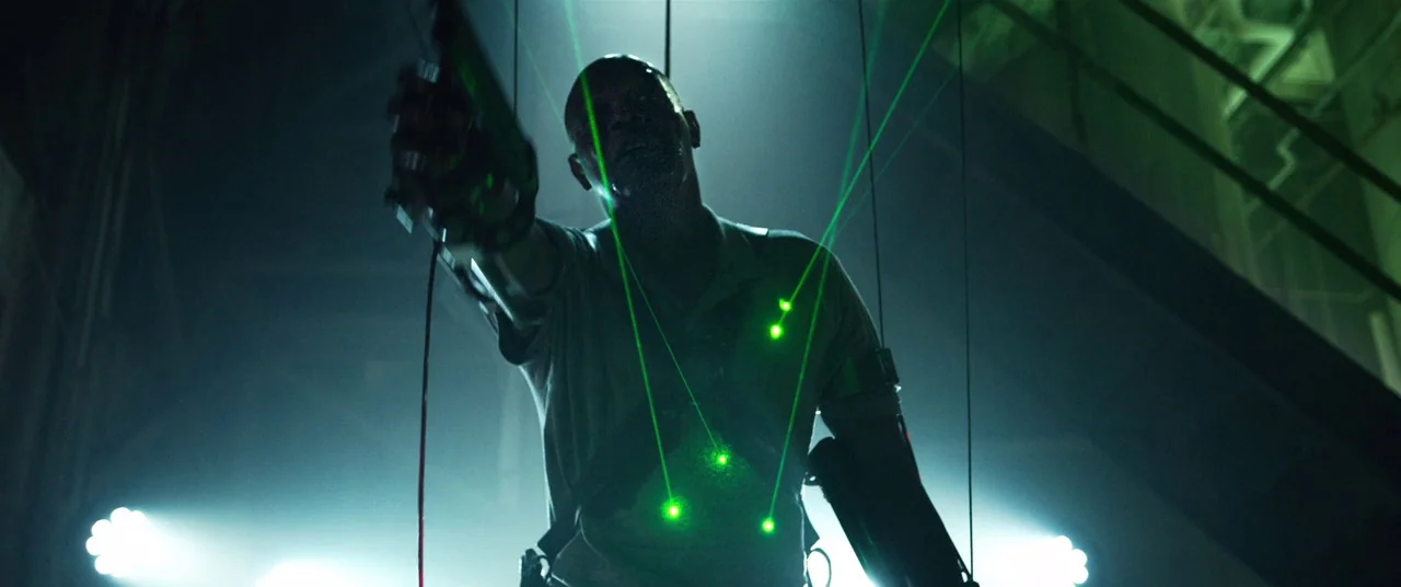 Spiral (2021) movie still: medium — A dark industrial space filled with hanging wires and metallic support structures. A man with a harness stands…