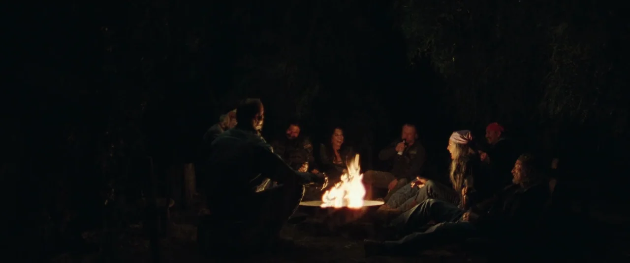 To Leslie (2022) movie still: wide — A group of people sits in a circle on dark ground around a campfire. Dense dark trees surround the clearing in…