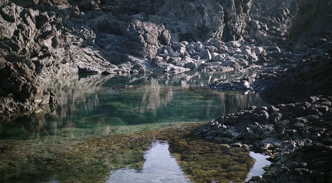 A Bigger Splash (2015) movie still: establishing — A dark rocky landscape features a clear pool of water trapped between jagged basalt. The surface…
