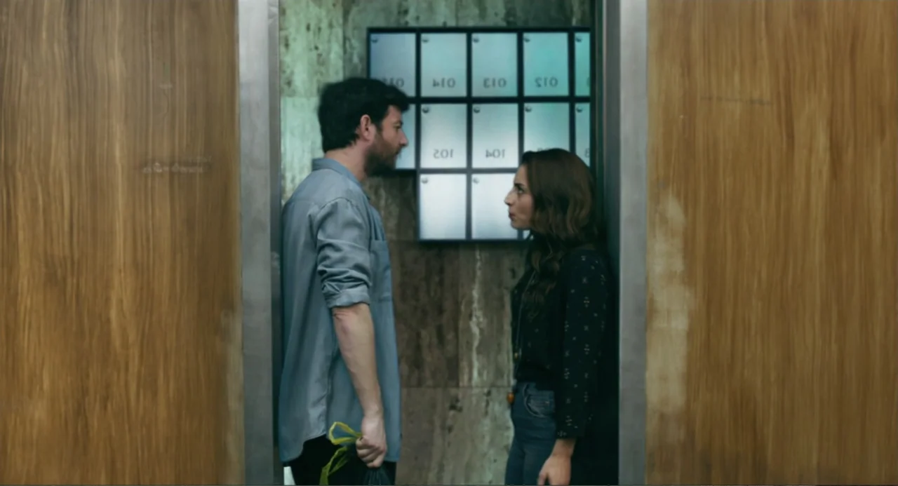 The Lift (2021) movie still: medium — A small mailroom with wood paneled walls and a wall of metal mail slots. A man and a woman stand facing each…