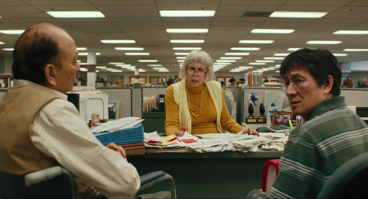Everything Everywhere All At Once (2022) movie still: medium — A cluttered administrative cubicle features piles of paperwork on a green desk…