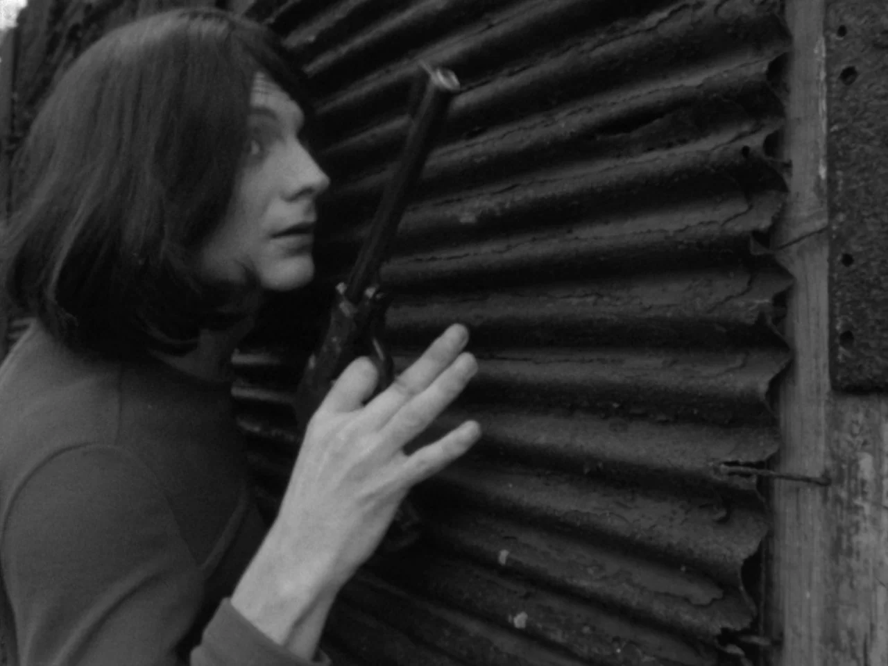 A Woman Kills (1968) movie still: medium — A person stands beside a wall of dark weathered corrugated metal sheeting. The individual grips a pistol…