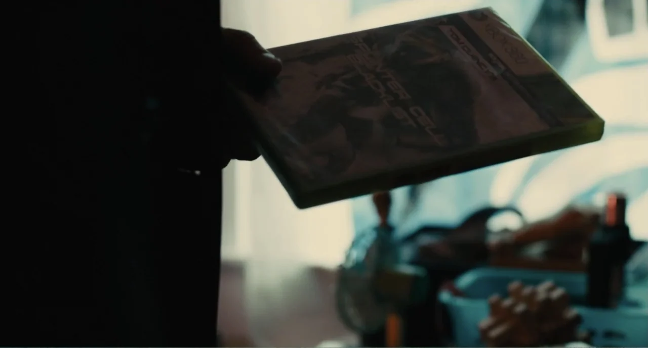 Lila & Eve (2015) movie still: closeup — An interior space contains miscellaneous cluttered items near a bright window. A hand holds a video game…