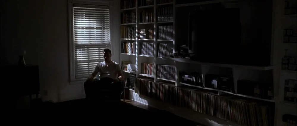 American Beauty (1999) movie still: wide — A dimly lit room features a large built-in white bookshelf filled with media cases. A man sits in a chair…