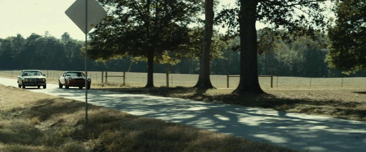The Case for Christ (2017) movie still: wide — A rural road lined with mature trees runs past a dry open field. Two vintage automobiles drive along…