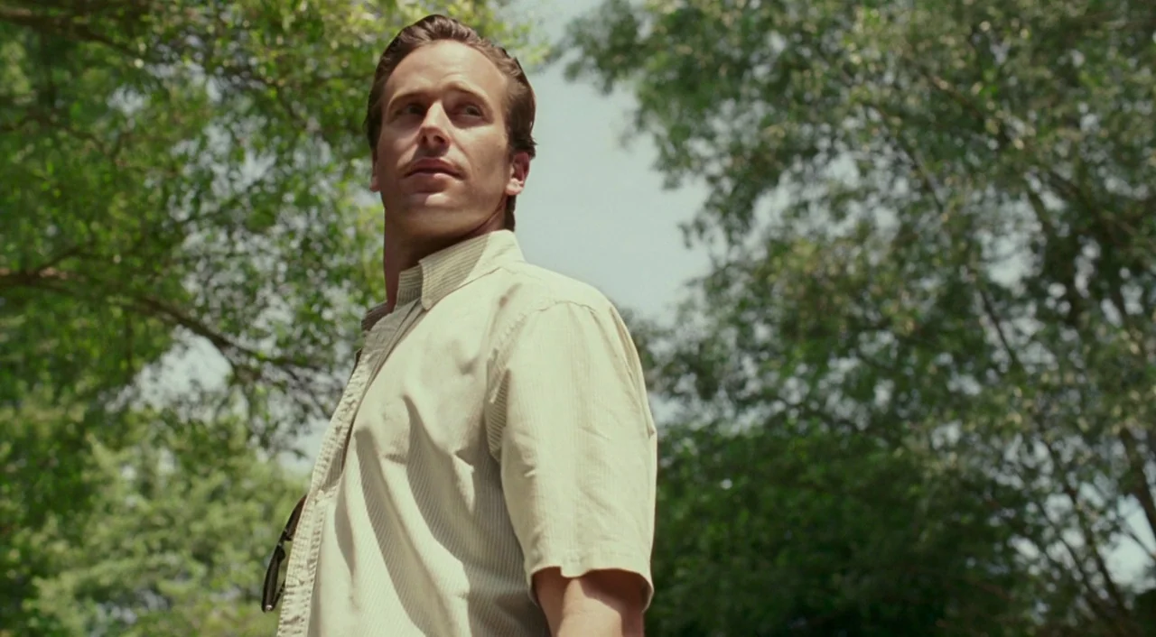 Call Me By Your Name (2017) movie still: medium — A man stands outdoors surrounded by dense green leafy trees during the day. He wears a…