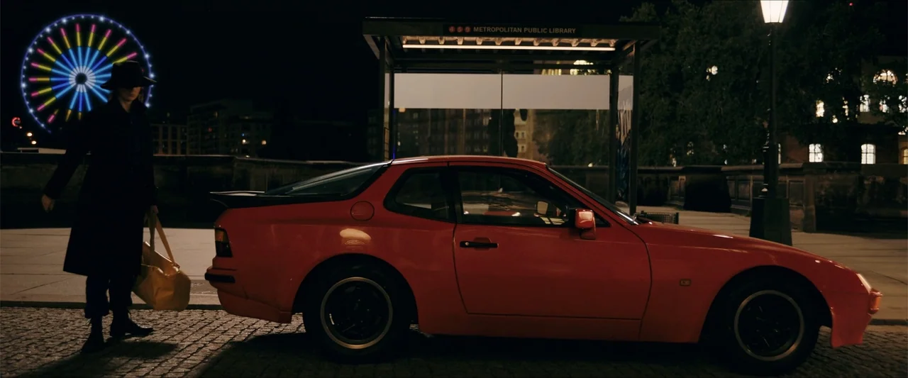 Gunpowder Milkshake (2021) movie still: wide — A night exterior showing a bus stop shelter and a bright red sports car. A person in a black coat…