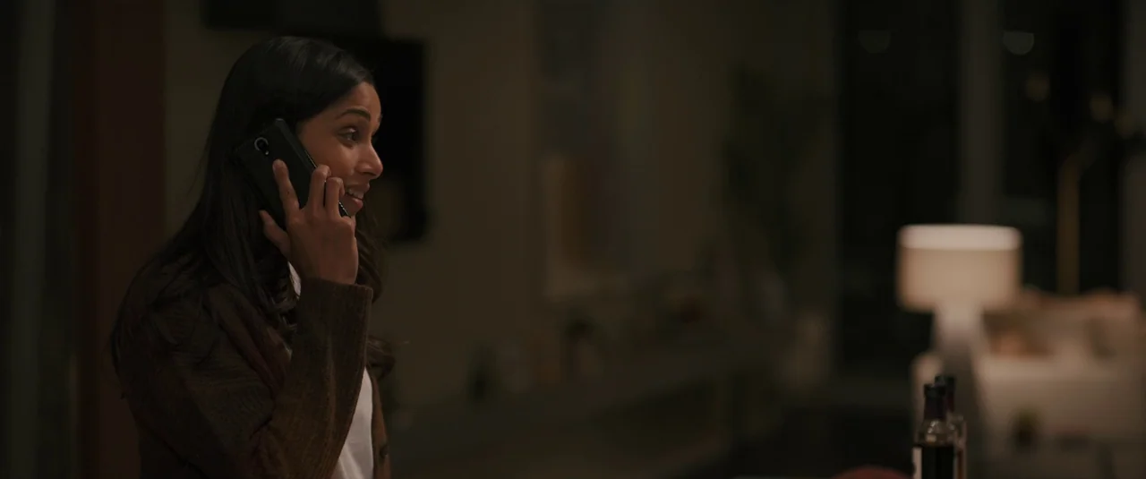 Intrusion (2021) movie still: medium — A dimly lit room in a modern home features dark furniture and soft lighting. A woman stands holding a phone…