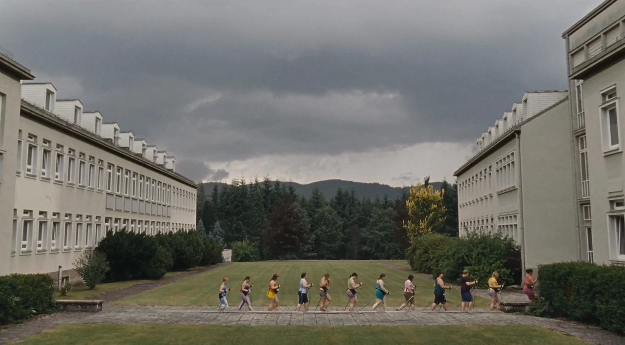 Paradise: Hope (2013) movie still: wide — A group of women in activewear walk in a line across a concrete pathway. Two identical buildings frame the…