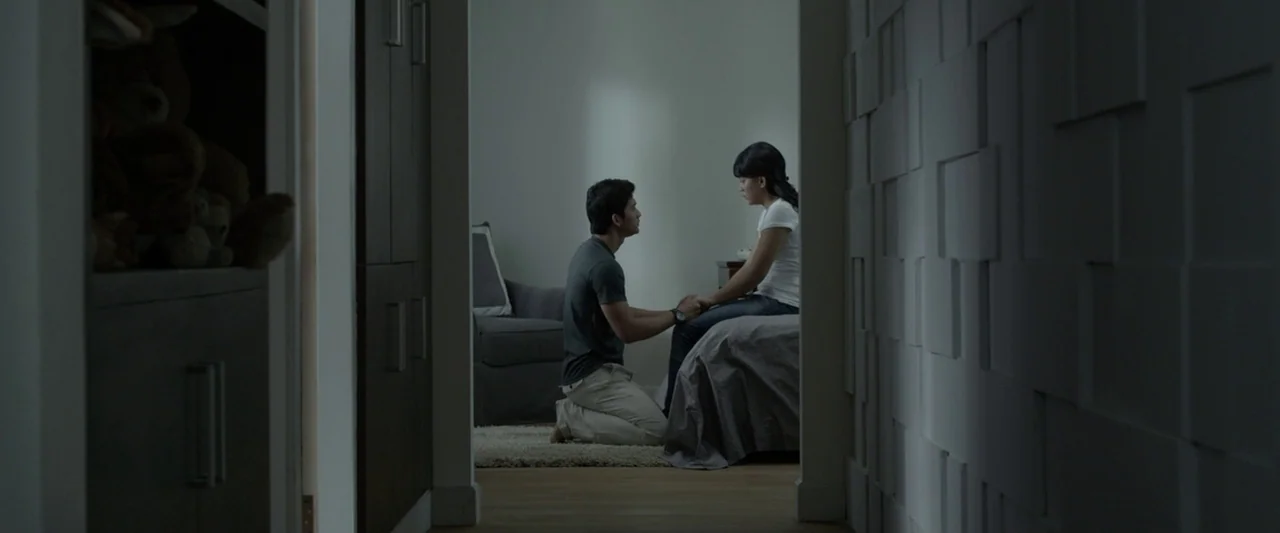 The Raid 2 (2014) movie still: wide — A dark hallway leads into a brightly lit room with a man kneeling before a woman. The couple sits facing each…