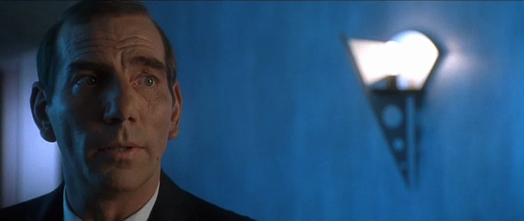 The Usual Suspects movie still: closeup — A dimly lit space features blue walls and a modern metallic wall sconce. An older man in a suit looks…