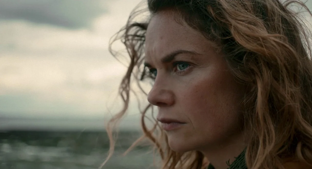Dark River (2017) movie still: closeup — A woman stands near the shore under a grey, overcast sky. The subject looks off-frame with a blank…