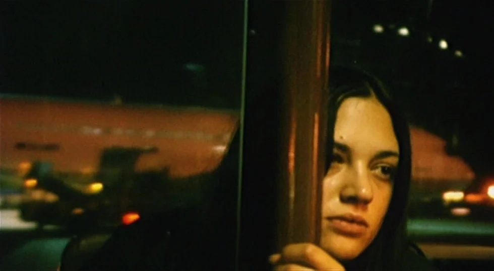 Scarlet Diva (2000) movie still: closeup — A dark bus interior at night shows a woman through a metal pole. Outside the window, blurry orange lights…