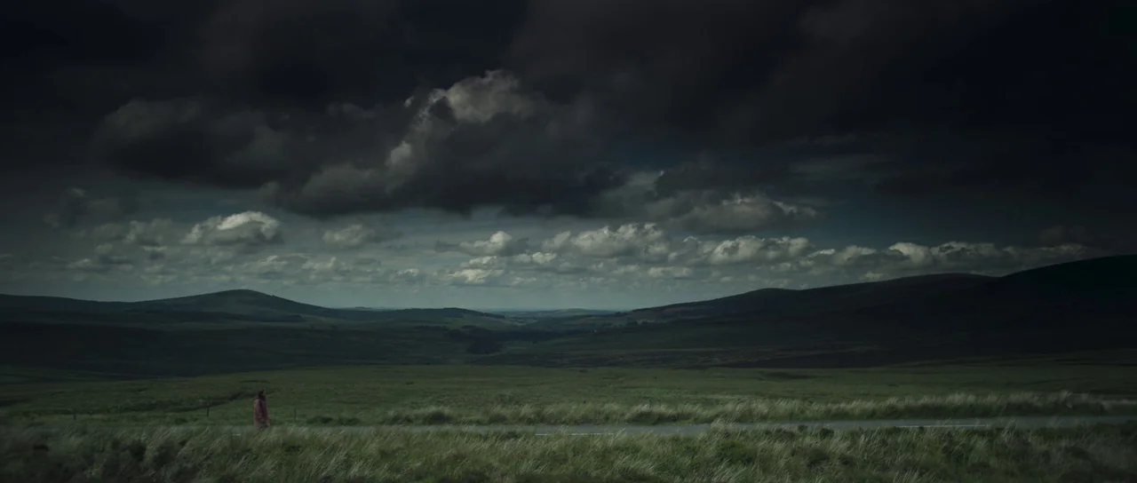 A Dark Song (2016) movie still: wide — A vast, rolling moorland landscape under a heavy, low-hanging overcast sky. A lone person in a red coat…
