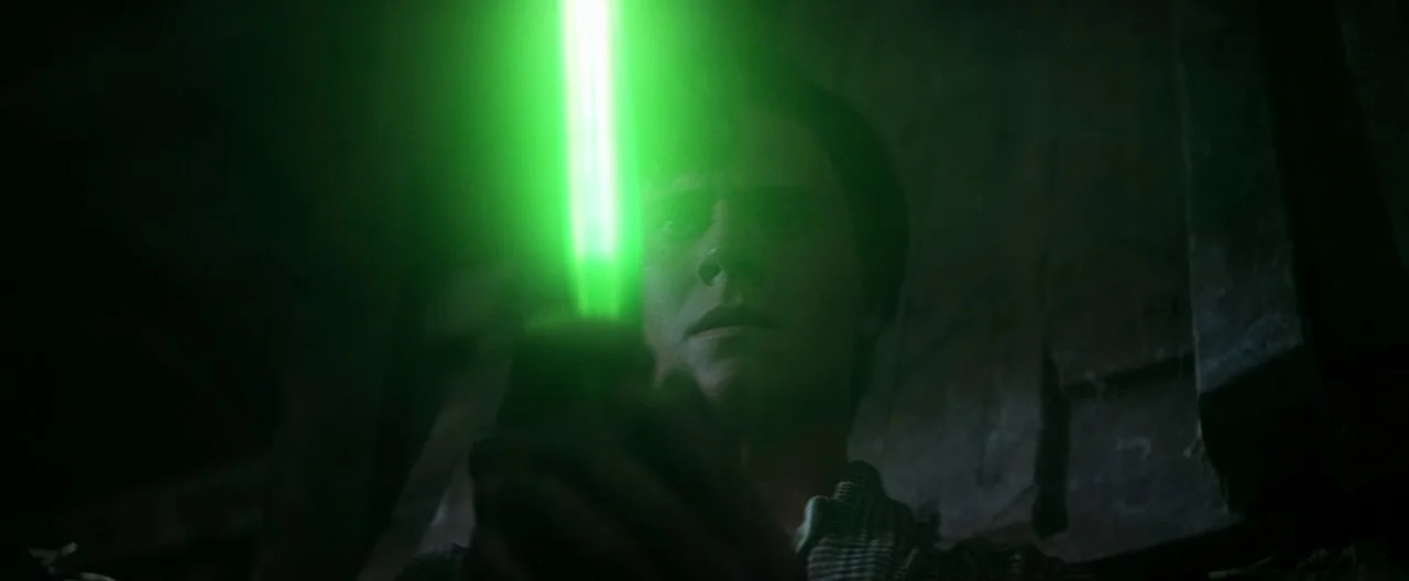 Superman (1978) movie still: closeup — A dark, textured interior space is illuminated by a single intense green light source. A young man holds the…