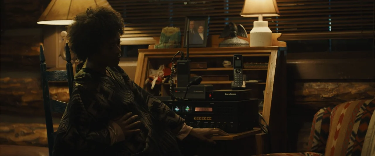 Those Who Wish Me Dead (2021) movie still: medium — A dimly lit log cabin interior features a wooden roll-top desk and radio equipment. A person in…