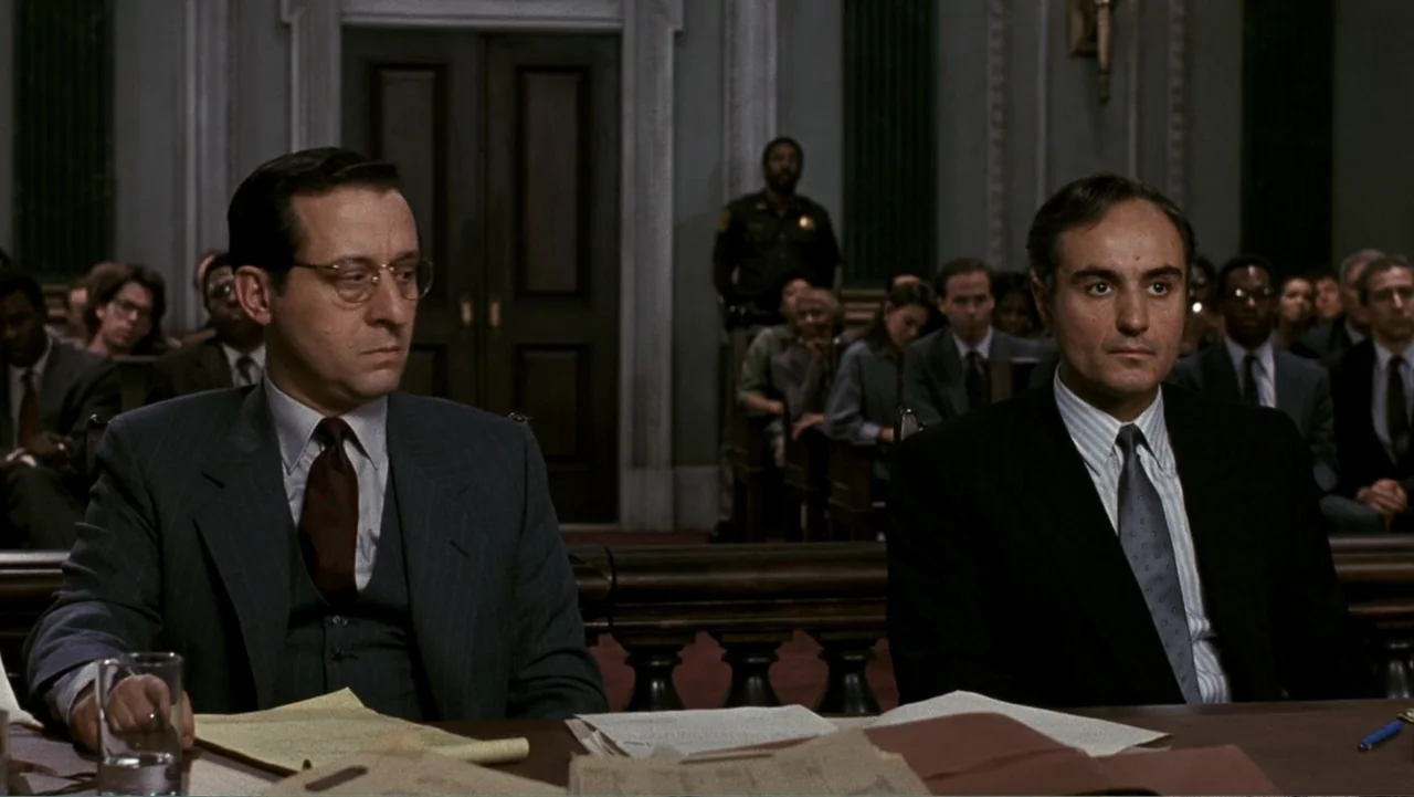 Presumed Innocent (1990) movie still: medium — A wood-paneled courtroom filled with seated spectators and a standing security officer behind them.…