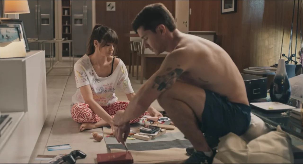 Proof of Love (2022) movie still: medium — A modern living room with wood paneled walls contains a young couple on the floor. The man and woman sit…