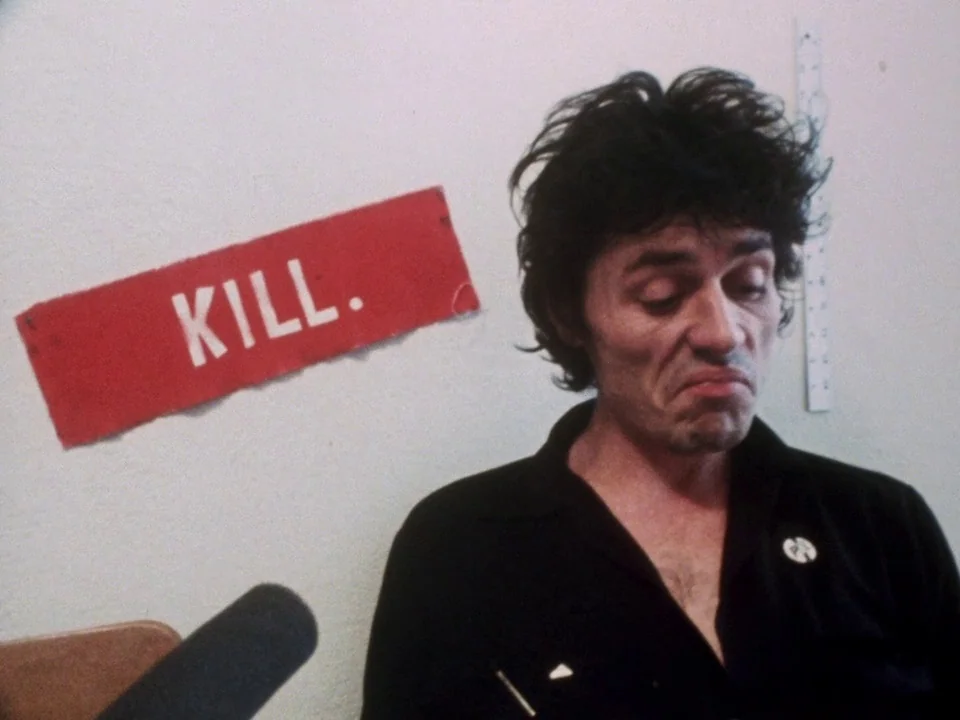 Decline of Western Civilization (1981) movie still: medium — A man stands before a blank white wall with a red sign reading KILL. The subject is…