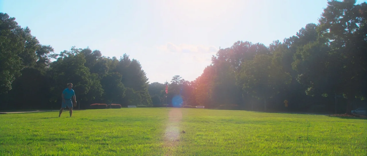 Greener Grass (2019) movie still: wide — A wide, grassy expanse is bordered by dense, mature trees under a bright sun. A man stands alone in the…