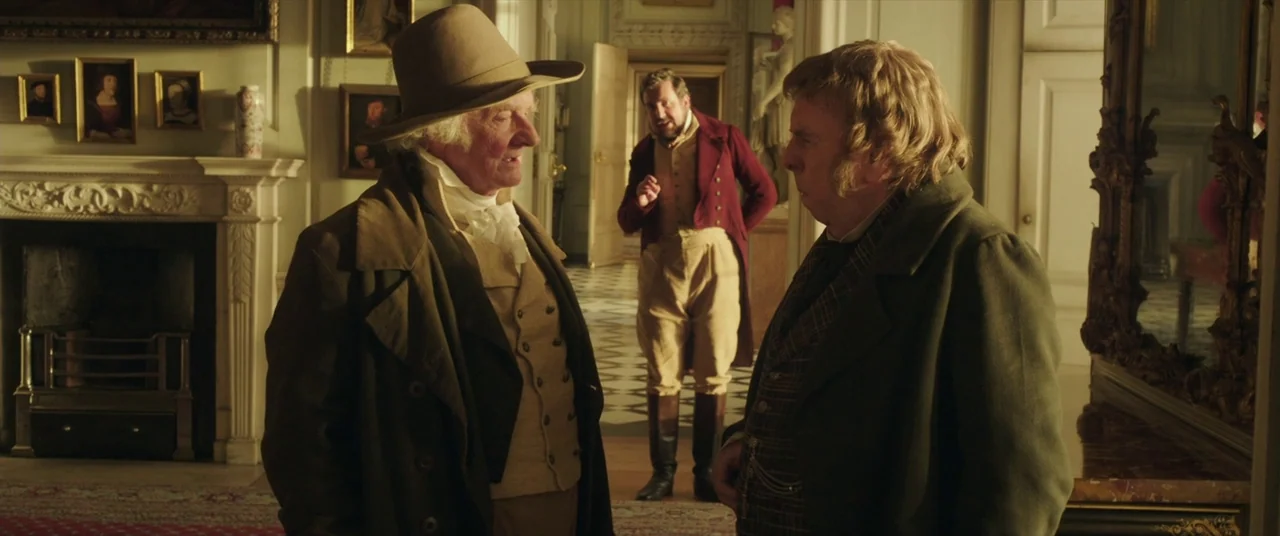Mr. Turner (2014) movie still: medium — An opulent room features a stone fireplace, hung portraits, and white wainscoted doors. Two men in period…