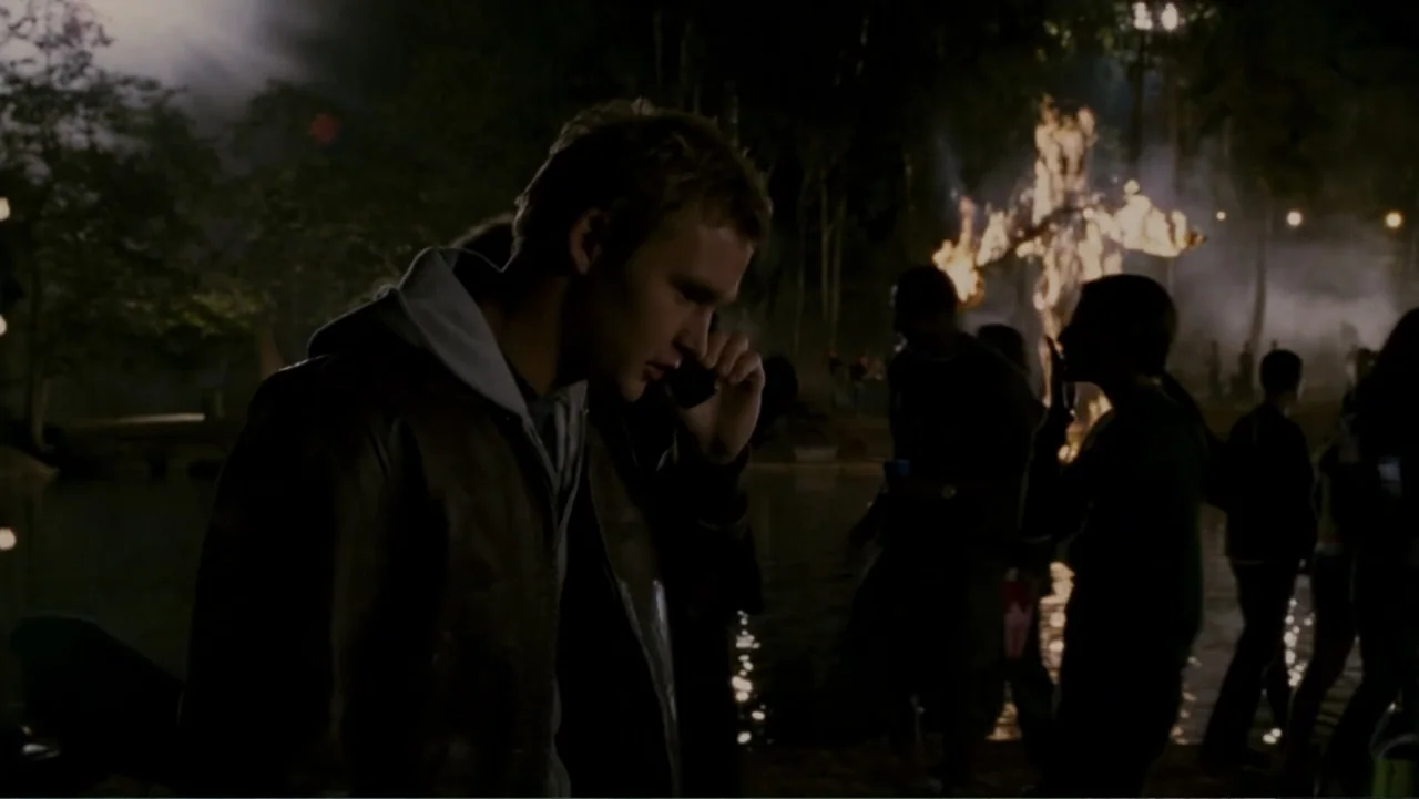 When a Stranger Calls (2006) movie still: medium — A night gathering by a dark lake featuring a bonfire burning in the background. A young man…