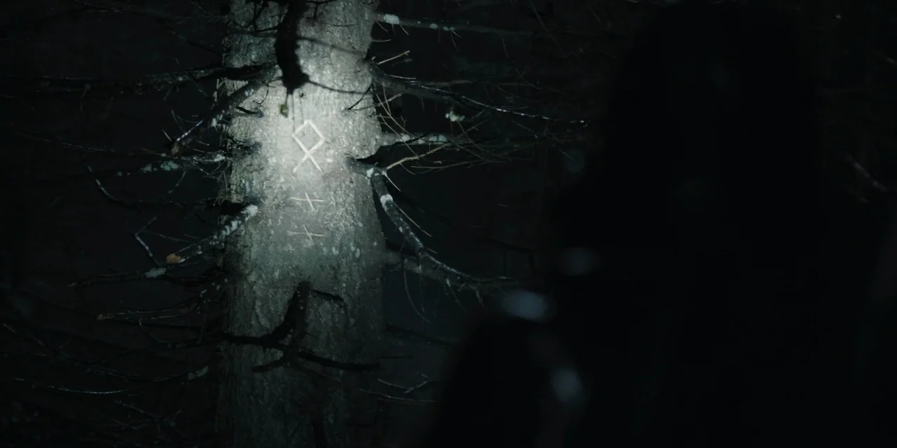 The Ritual (2017) movie still: medium — A dark, dense woodland environment features a tree trunk with carved runic symbols. A person obscured in…