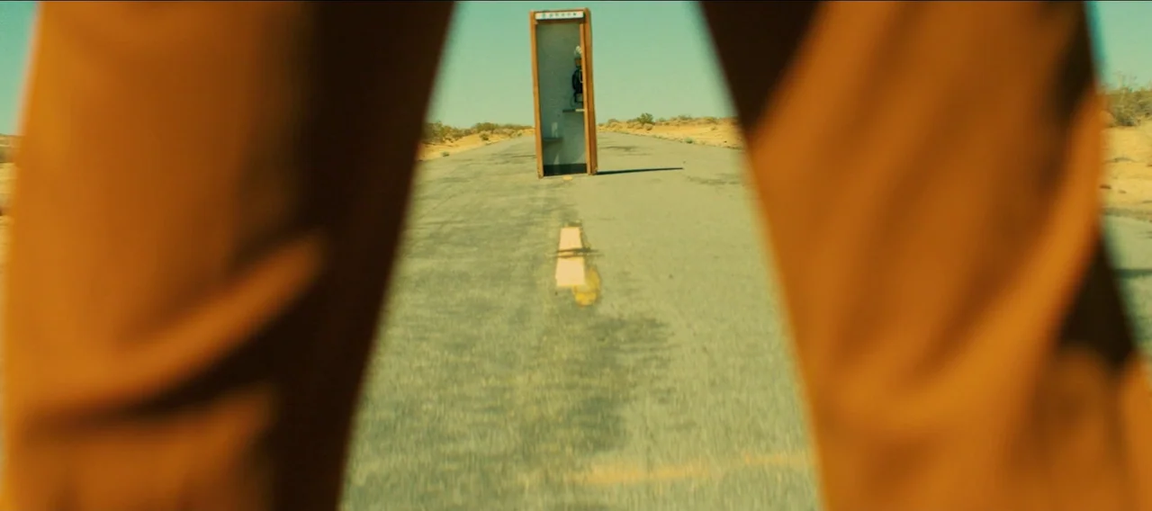 Fuzzy Head (2023) movie still: wide — A remote, paved road stretches through a flat, arid desert landscape under clear skies. A vintage outdoor…
