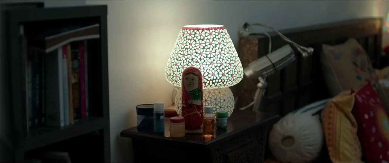 Cheekatilo (2026) movie still: medium — A bedroom side table sits next to a shelf filled with various books. A decorative lamp glows beside a…