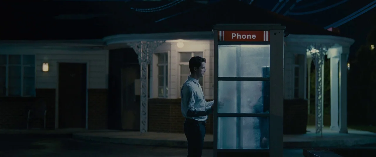 Stoker (2013) movie still: wide — A dark night exterior features a motel facade with vintage architectural detailing and white columns. A man stands…
