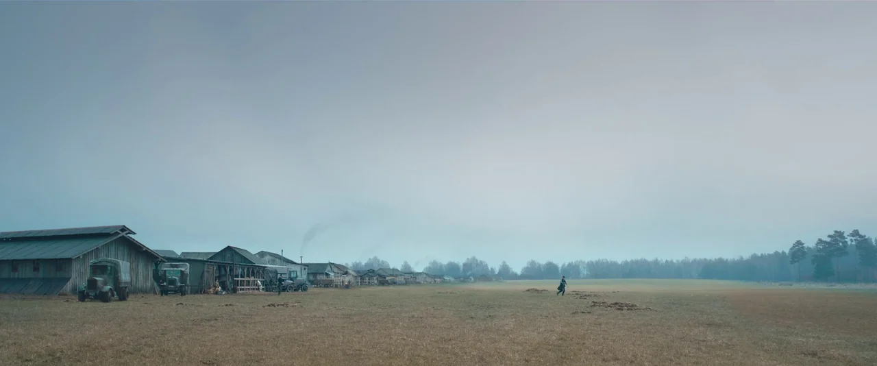 All Quiet on the Western Front (2022) movie still: wide — An expansive, flat, and sparse field features a line of rustic wooden buildings. A lone…