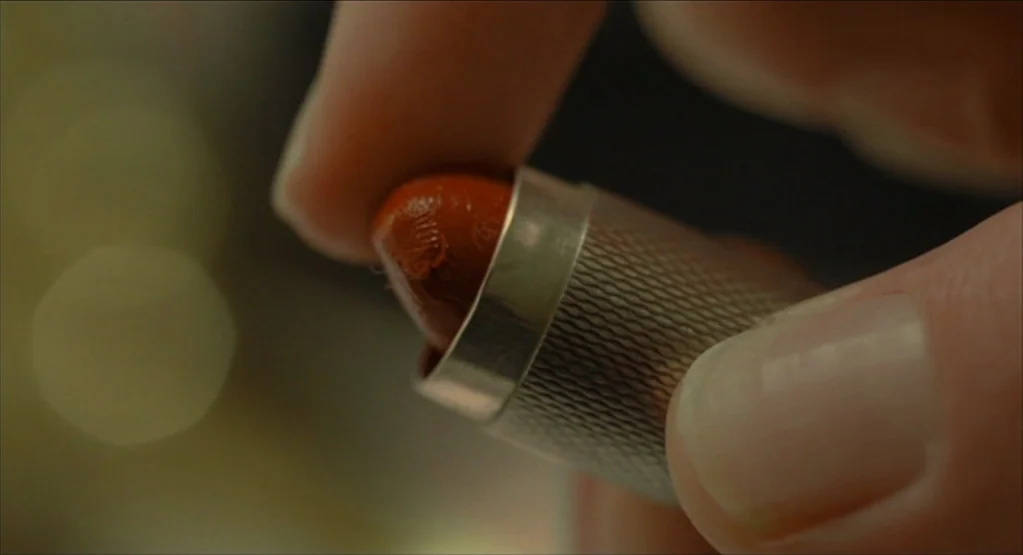 The Fall movie still: closeup — A close-up view shows a finger touching a small dollop of orange lipstick. The lipstick is contained within a silver…