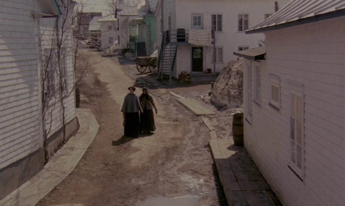 Kamouraska (1973) movie still: wide — A dirt road passes between white clapboard houses in a rural nineteenth-century settlement. Two women walk…