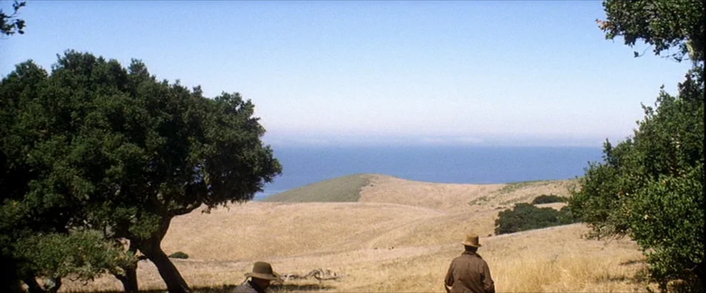 There Will Be Blood (2007) movie still: wide — Coastal hills covered in dry grass descend toward the Pacific Ocean under a clear sky. Two men in…