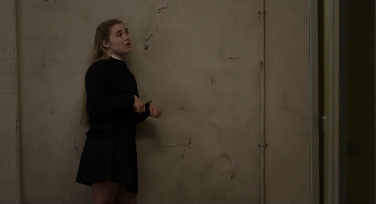 The Falling movie still: medium — A dimly lit interior space features a weathered wall with peeling off-white paint. A young woman stands against…