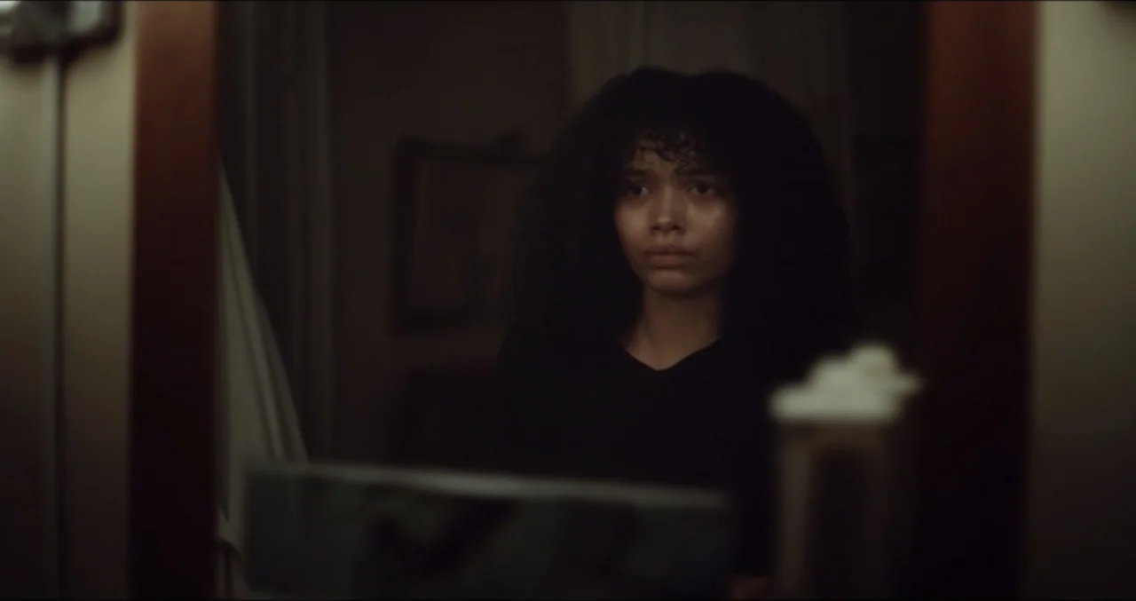 Eye for an Eye (2025) movie still: medium — A dimly lit room containing a wall-mounted mirror above a vanity. A woman with dark curly hair stares…